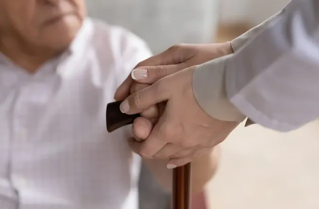 Ein Mann mit Gehstock wird von einem Physiotherapeuten im Kittel während eines Hausbesuchs geführt, um Unterstützung bei der Mobilität zu erhalten.